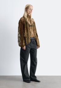 Durchsichtige, olivgrüne Spitzbluse mit hohem Kragen, langen Ärmeln und floralem Muster, kombiniert mit hochtaillierten, lockeren schwarzen Jeans und spitzen Ballerinas.