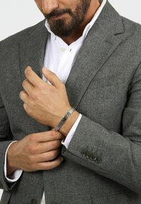 Bracciale d'argento con motivi geometrici intrecciati indossato a un polso, abbinato a un blazer grigio con texture e una camicia bianca con colletto abbottonato.