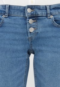 Jean en denim bleu avec trois boutons métalliques, poches avant, passants de ceinture et coutures visibles, porté avec un haut blanc.