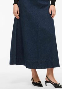 Długa spódnica jeansowa w ciemnym niebieskim kolorze o rozkloszowanym kroju. Charakteryzuje się czystym wykończeniem i delikatną teksturą. Połączona z czarnymi sandałkami na obcasie z paskiem z tyłu.