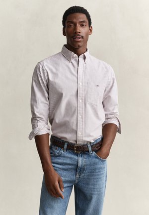 Mann mit kurzen lockigen Haaren, der ein weißes und hellrosa gestreiftes Hemd mit Knöpfen und blaue Jeans mit einem braunen geflochtenen Gürtel trägt.