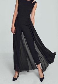 Frau in einem ärmellosen schwarzen Jumpsuit mit transparenten langen Überlagerschalen und schwarzen spitzen High Heels, stehend vor einem grauen Hintergrund.