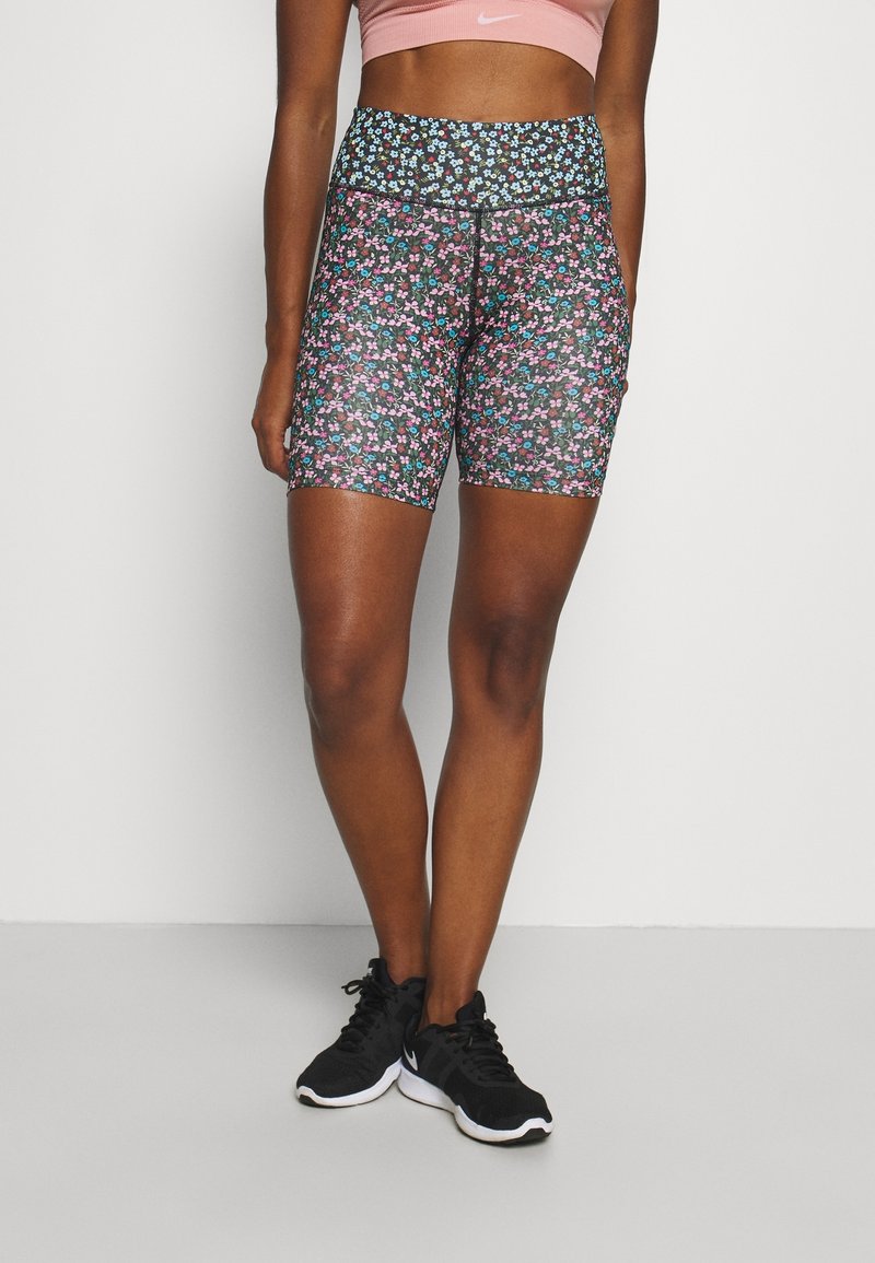 Femme portant un short de vélo taille haute à imprimé floral, associé à des chaussures de course noires et un soutien-gorge de sport rose sur un fond uni.