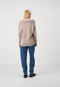 Pull beige oversize avec un large col, tricot texturé et ourlet côtelé, associé à un jean bleu ample et des chaussures noires épaisses.