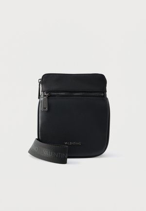 Sac bandoulière noir Valentino avec poche zippée à l'avant et sangle ajustable avec logo sur fond blanc.