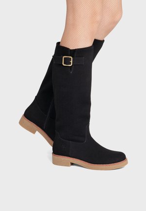 Jambes portant des bottes noires montantes avec semelles beiges et détail de boucle dorée, associées à des bas résille beiges.
