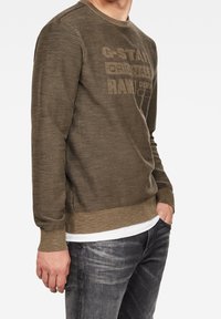 Man i brun G-Star Raw sweatshirt, vit undertröja och blekta svarta jeans med händerna i fickorna, mot vit bakgrund.