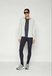 Femme portant des lunettes de soleil noires, une veste zippée gris clair, des leggings noirs avec poches et des baskets gris clair, se tenant devant un fond uni.
