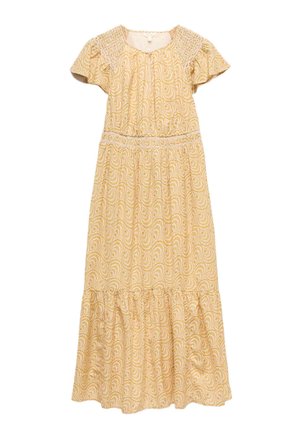 Frau trägt ein gelbes, gemustertes Midikleid, eine braune Wildlederweste und braune Schnürsandalen und steht mit entspannt an den Seiten hängenden Armen.