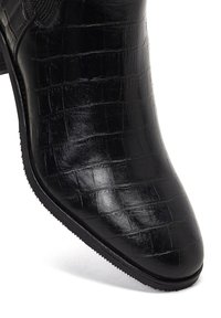 Botín negro de imitación de piel con textura de cocodrilo, puntera afilada y tacón bajo. Incluye paneles elásticos para facilitar su uso.