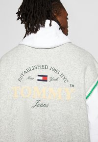 Graues Fleece-Sweatshirt mit weißem Kapuzenfutter. Mit besticktem grünen und gelben Schriftzug: "TOMMY Jeans" und Logo. Akzente mit grünen Streifen.