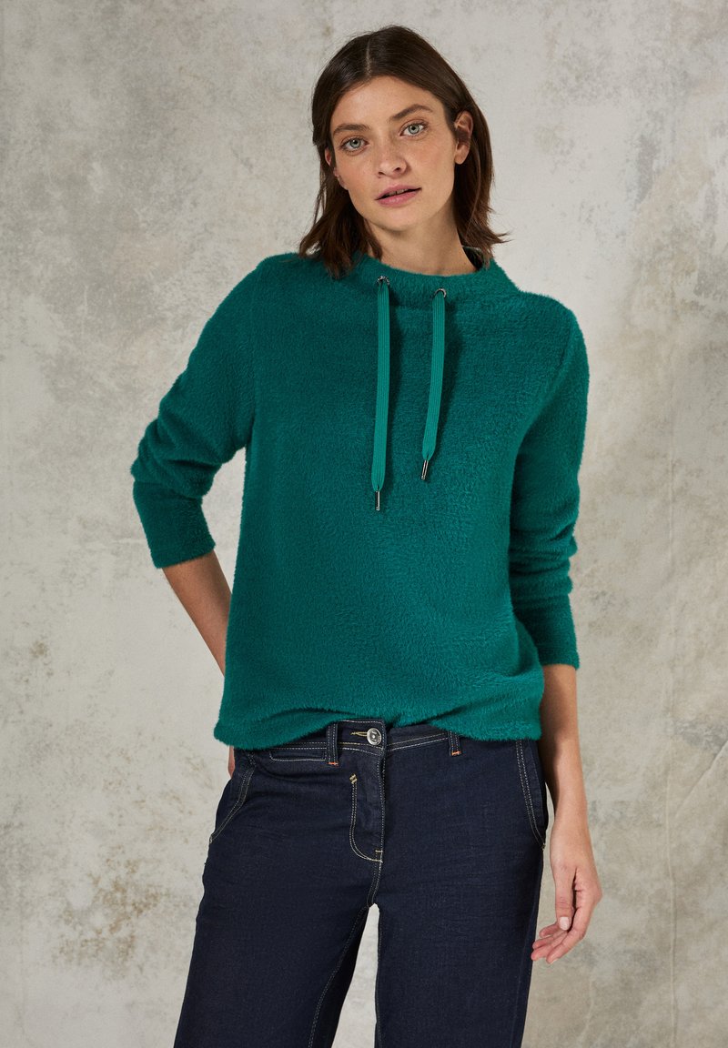 Türkisfarbener, flauschiger Pullover mit hohem Kragen und Kordelzügen, kombiniert mit dunkler Jeans. Weiche Textur, lockere Passform und Dreiviertelärmel.