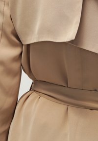 Beżowa sukienka z miękkiego, drapowanego materiału, z paskiem w talii i plisowanymi detalami u dołu. Gładka tekstura z delikatnym połyskiem.