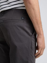 Homme portant un pantalon gris foncé et une chemise rayée noir et blanc, la main posée près de la poche latérale sur un fond clair.