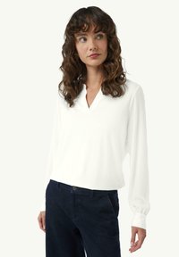 Weißes langärmeliges Shirt mit V-Ausschnitt, aus weichem Stoff gefertigt. Die Manschetten sind leicht gerafft und werden mit dunklen Hosen kombiniert.