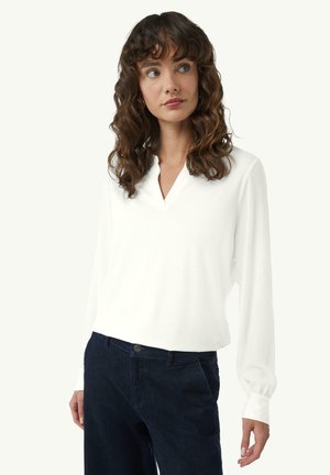 Weißes langärmeliges Shirt mit V-Ausschnitt, aus weichem Stoff gefertigt. Die Manschetten sind leicht gerafft und werden mit dunklen Hosen kombiniert.