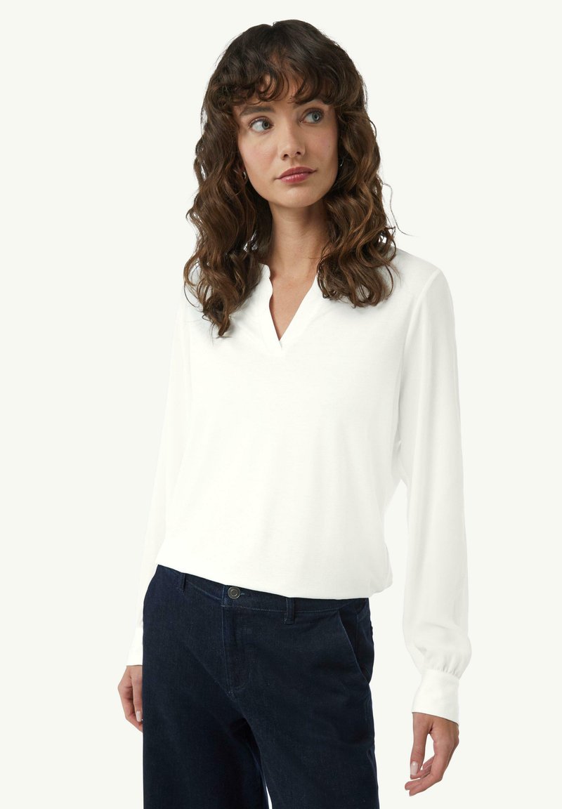 Weißes langärmeliges Shirt mit V-Ausschnitt, aus weichem Stoff gefertigt. Die Manschetten sind leicht gerafft und werden mit dunklen Hosen kombiniert.