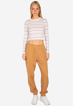 Young woman standing, wearing a white striped long-sleeve crop top, tan elastic waist joggers, and white sneakers, on a plain background.