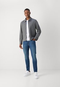 Siva zip-up jakna preko bijele majice, nosena s plavim uskim trapericama i bijelim tenisicama. Jakna ima strukturirani ovratnik i suptilnu teksturu.