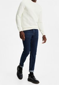Vit texturerad tröja med ett ribbat mönster, kombinerad med mörkblå slim-fit jeans och svarta snörskor.