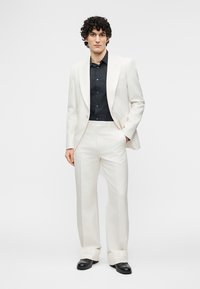 Άνδρας στέκεται φορώντας λευκό κοστούμι με μαύρο πουκάμισο και μαύρα παπούτσια, με το χέρι στην τσέπη, μπροστά σε απλό λευκό φόντο.