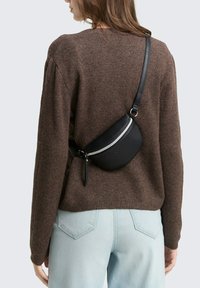 Marsupio in pelle nera con cerniera argentata lucida, indossato in diagonale sulla schiena, abbinato a un maglione di maglia marrone e jeans azzurri chiari.