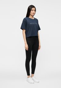 T-shirt curta azul-marinho com a inscrição "MICHAEL KORS", combinada com leggings pretas e ténis brancos, sobre um fundo simples.