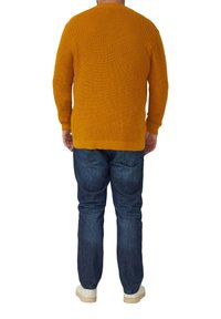 Kardigan w kolorze musztardowej żółci, z długimi rękawami, ściągaczami na mankietach i u dołu. Noszony z niebieskimi dżinsami i białymi butami, widziany od tyłu.