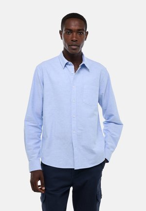 Homme portant une chemise bleu clair à boutons avec une poche poitrine et un pantalon foncé, debout devant un fond clair uni.