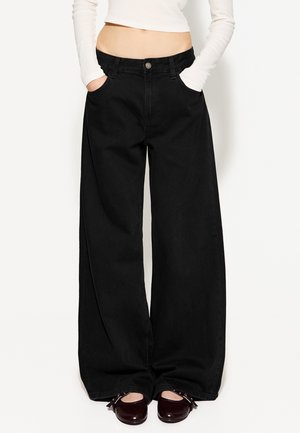 Weite schwarze Jeans mit Fronttaschen und Knopfverschluss. Glatte Textur und lockerer Schnitt. Kombiniert mit dunklen, runden Schuhen.