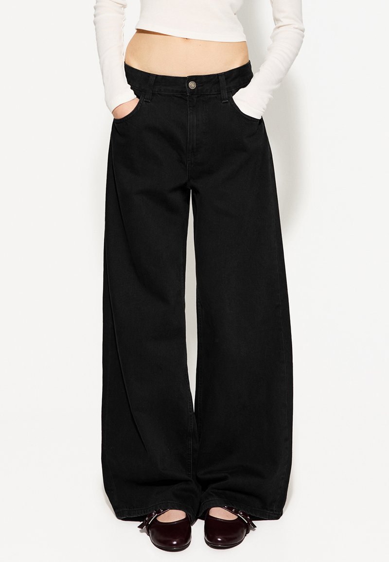Wide-leg black denim pants with front pockets and a button closure. Smooth texture and relaxed fit. Paired with dark, rounded shoes.