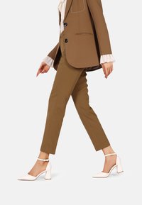 Scarpe con tacco alto bianche, con punta affusolata e cinturino alla caviglia, abbinate a pantaloni marroni aderenti e un blazer abbinato. Texture liscia, design minimale.