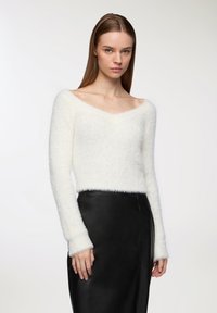 Maglione bianco morbido con scollatura a V, maniche lunghe e polsini a coste, abbinato a una gonna in pelle nera, creando un contrasto di texture delicato.