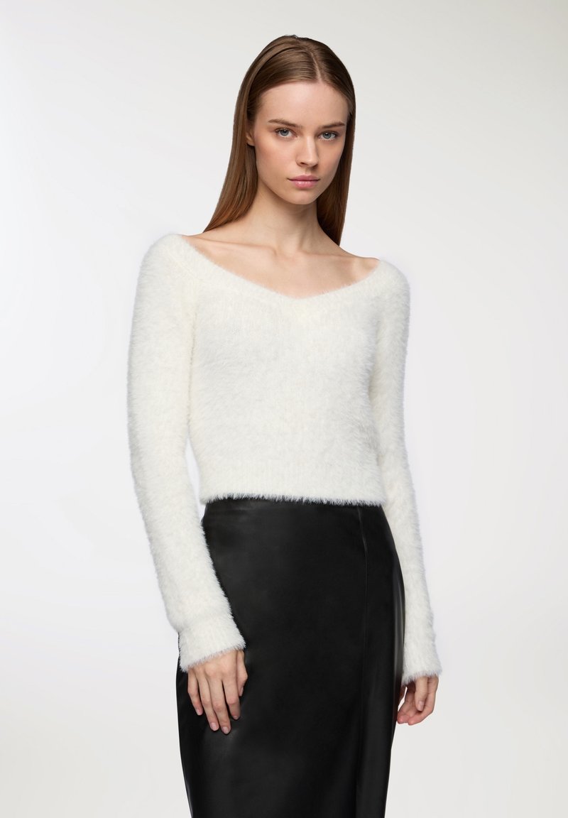 Maglione bianco morbido con scollatura a V, maniche lunghe e polsini a coste, abbinato a una gonna in pelle nera, creando un contrasto di texture delicato.