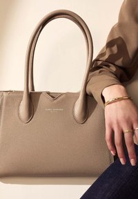 Beige leren tote bag met textuur, voorzien van twee ronde handvatten, een goudkleurig logo en een ritsluiting. Hand gedeeltelijk zichtbaar, met ringen aan.