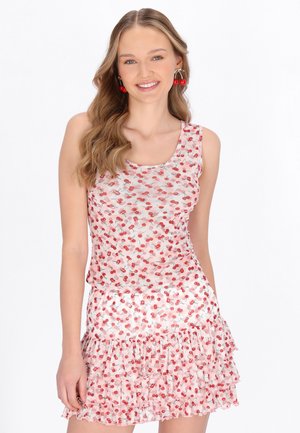 Mosolygó nő hullámos hajjal, cseresznye mintás ujjatlan ruhában és hozzáillő cseresznye alakú fülbevalóval, fehér háttér előtt.