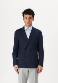 Navy double-breasted blazer in lightweight fabric, featuring two front pockets, pointed lapels, and three black buttons, worn over a light striped shirt.