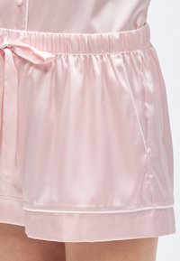 Ροζ σατέν σορτς με ελαστική μέση, διακοσμημένα με φιόγκο και πλαϊνές τσέπες. Το ύφασμα έχει ομαλή, γυαλιστερή υφή.