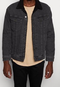 Svart denimjacka med en strukturerad svart fleecekrage, knäppning framtill och fickor; kombinerad med en beige t-shirt och svarta byxor.