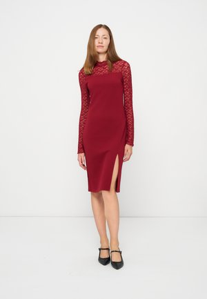 Femme debout sur fond blanc, portant une robe bordeaux à manches longues en dentelle avec une fente sur la cuisse, associée à des chaussures Mary Jane noires.