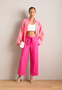 Rosa, overdimensjonert skjorte med knapper, hvit crop-top, lyserosa vide bukser og beige høyhælte sandaler med stropper. Holder en liten hvit veske.