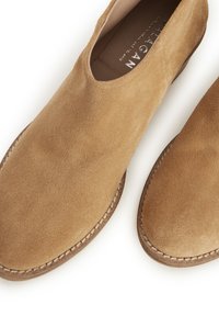 Zapatos slip-on de ante en color marrón claro con puntera redondeada, detalle de costura alrededor de la suela y forro interior de cuero suave.