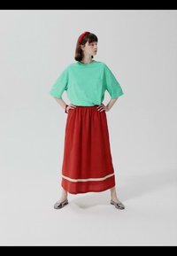 Femme debout, mains sur les hanches, portant un tee-shirt oversize vert menthe, une longue jupe rouge avec un ourlet blanc, des ballerines argentées et un bandeau rouge.
