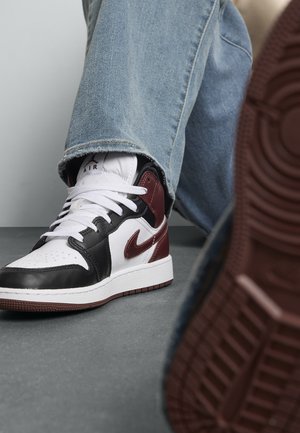 Personne portant un jean bleu et des sneakers Nike Air blancs, noirs et bordeaux, un pied sur une surface grise, montrant le motif de la semelle de la chaussure.