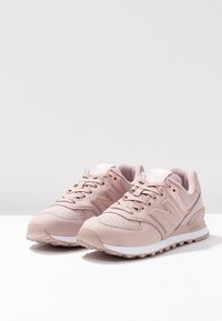 Vaaleanpunaiset urheilukengät, joissa on verkko- ja keinonahkamateriaaleja, rei'itettyjä yksityiskohtia ja valkoinen pohja. Sivulla on suuri "N"-logo.