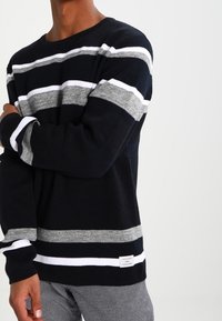 Maglione a righe nere e grigie con dettagli bianchi, texture a coste, colletto girocollo e etichetta del marchio in fondo, indossato con pantaloni grigi.
