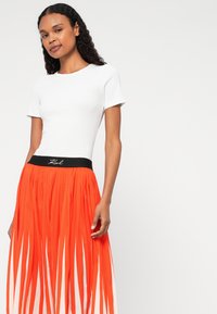 Fehér, rövid ujjú póló, amelyet egy hosszú, narancssárga redőzött szoknyával párosítottak, fekete derékkal, amelyen "Karl" logó található. Simbeszövésű anyag élénk színekkel.