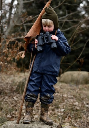 Lapsi, jolla on laivaston sininen vedenpitävä takki ja housut, pitää mustia kiikareita kädessään ja käyttää vihreää hattua, seisoo kivellä ja jalassaan mutaiset kukkakuvioiset saappaat.