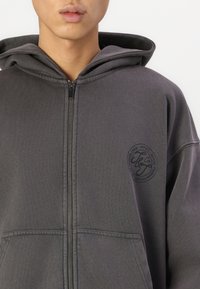 Hoodie zippé gris en coton avec capuche, présentant un patch logo rond, deux poches avant et des poignets côtelés pour un ajustement sûr.
