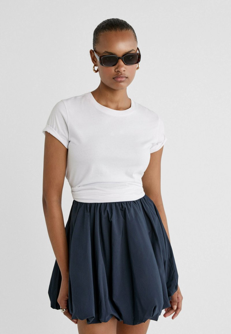 T-shirt bianco aderente con maniche arrotolate abbinato a una gonna voluminoso blu navy. Gli accessori includono occhiali da sole neri e orecchini a cerchio.